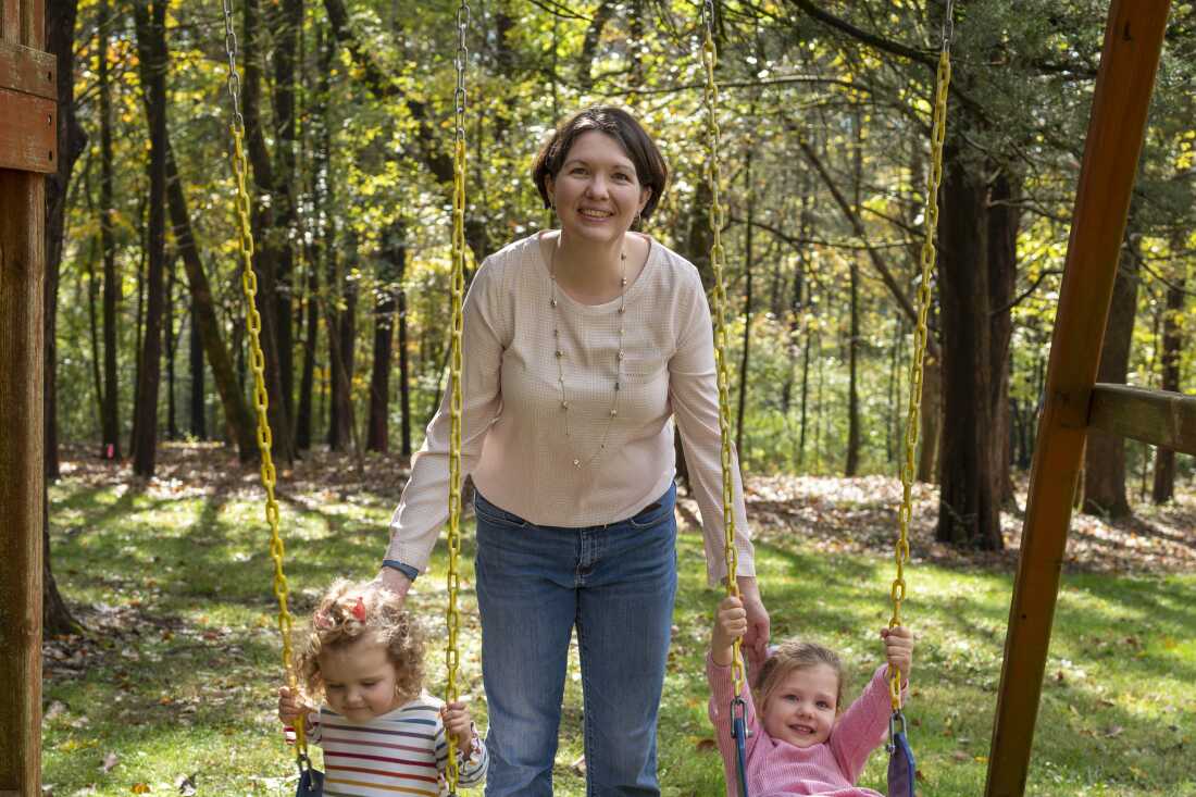 Laura Terry stands in between her two daughters, who are seated on a swing set.