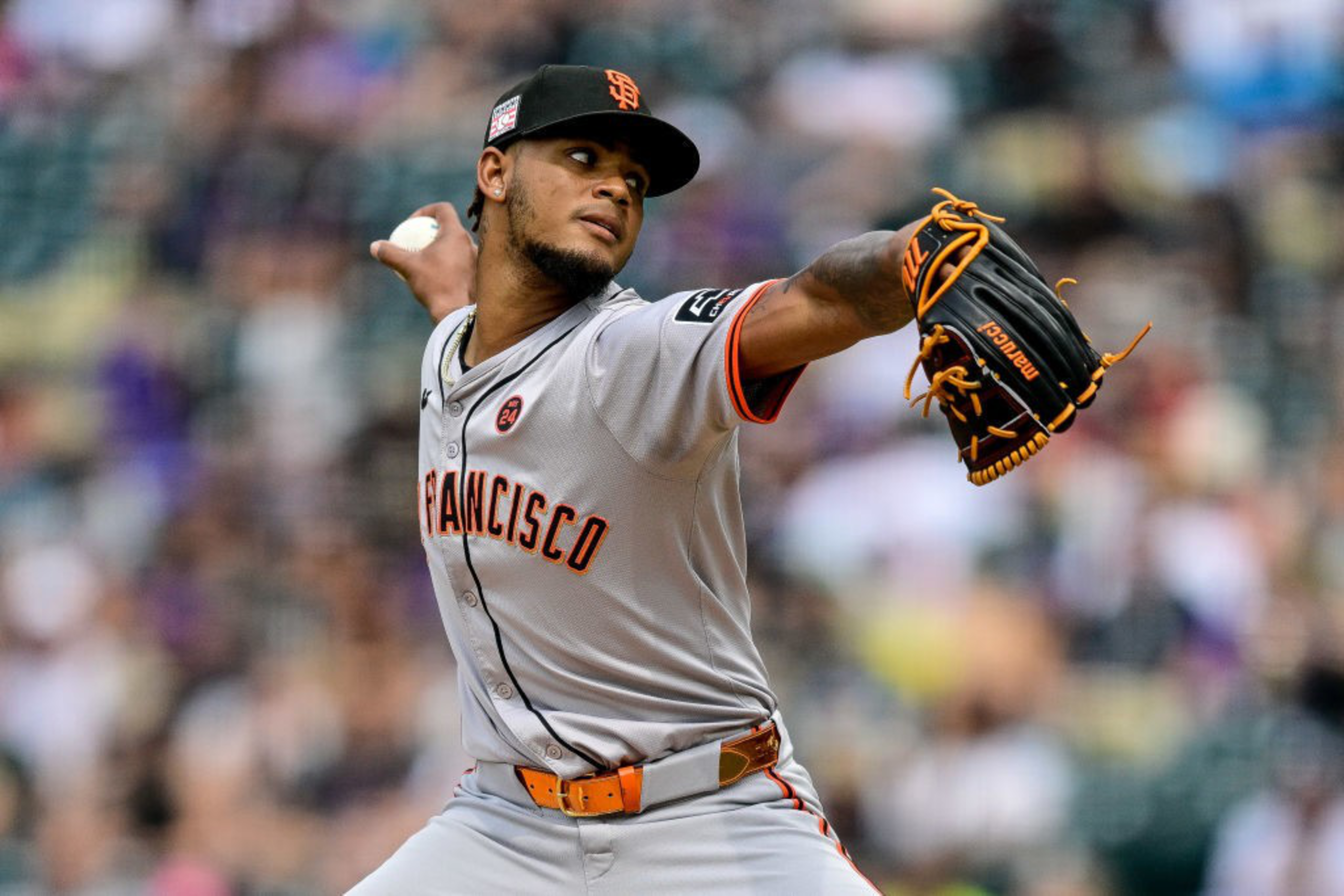 A baseball player in a gray uniform with “San Francisco” across the chest is pitching, wearing a black cap and glove, with a blurred stadium background.