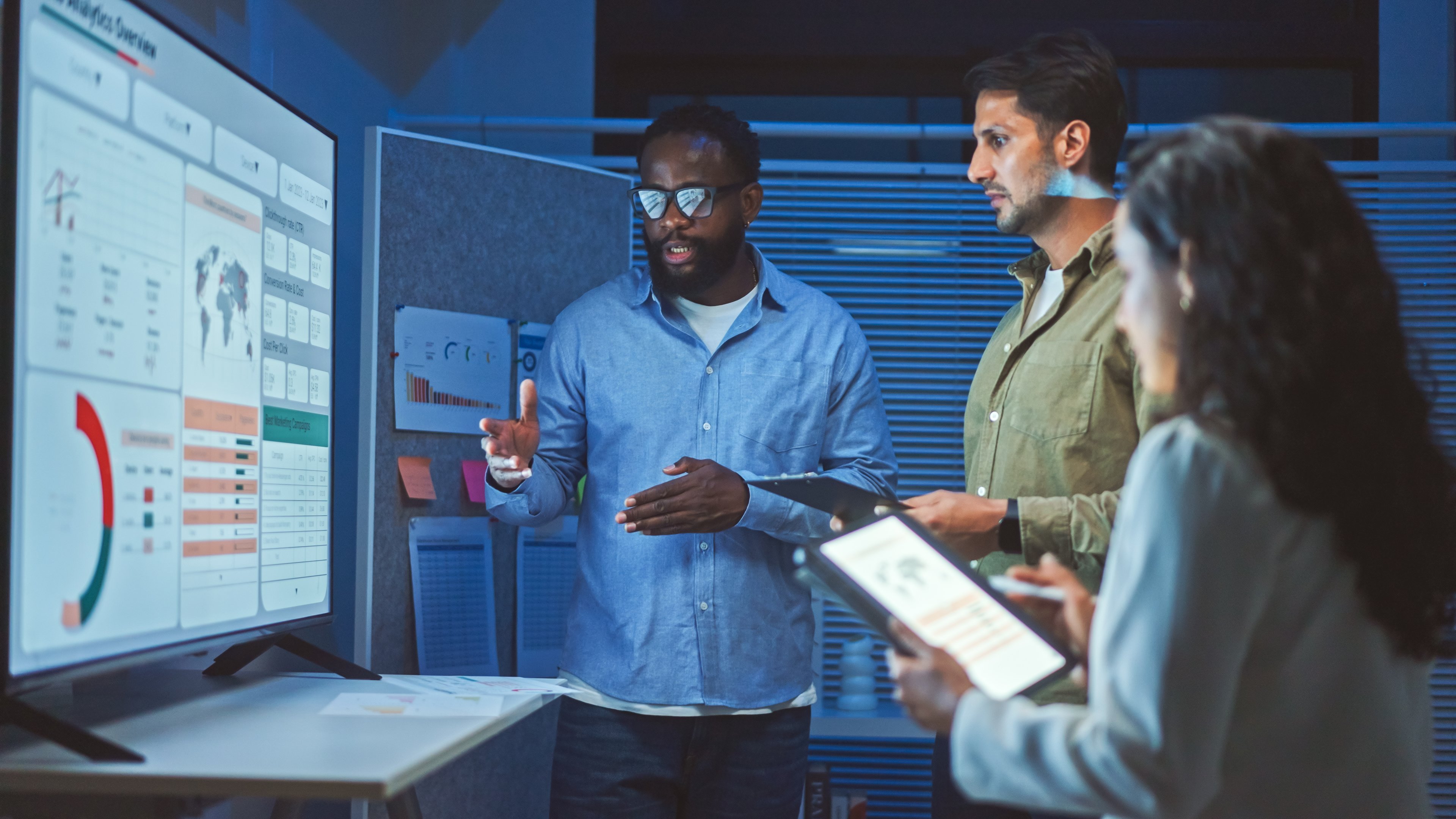 Three people standing in front of a monitor and discussing a chart on the screen.