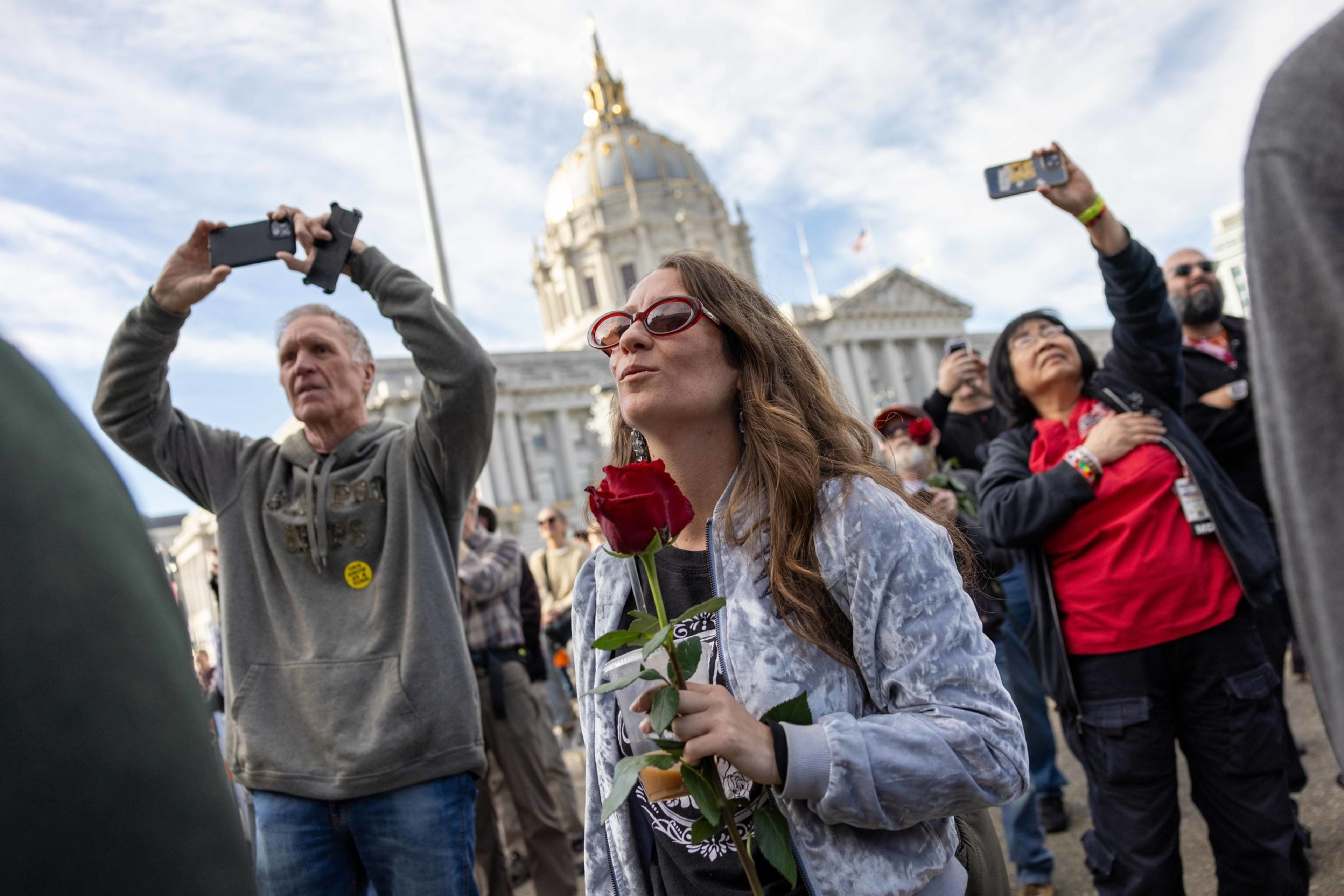 A woman holding a red rose stands among a crowd of people taking photos, with a grand building featuring a gold-topped dome in the background.
