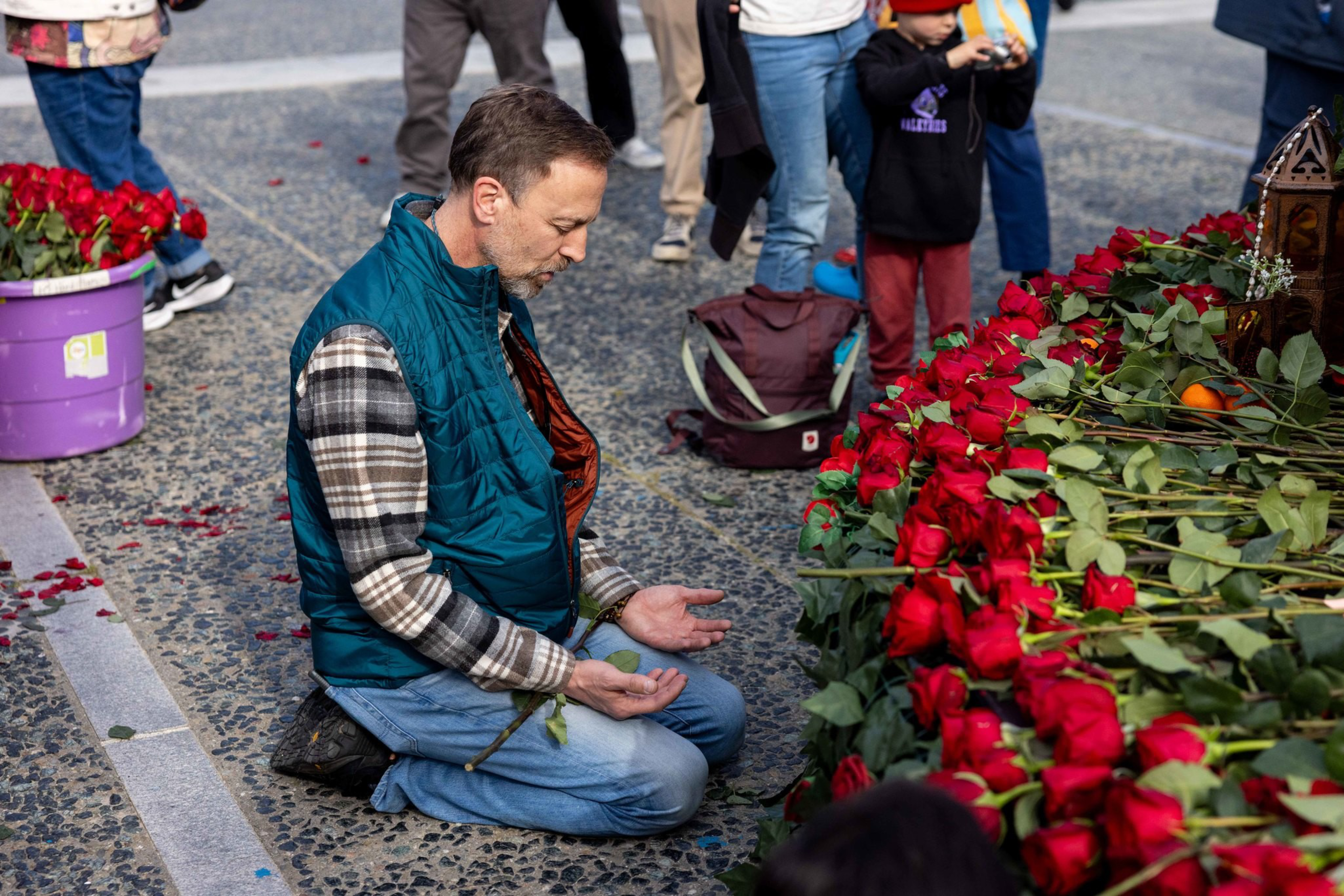 A man kneels solemnly beside a large pile of red roses, holding one rose and surrounded by people standing nearby.