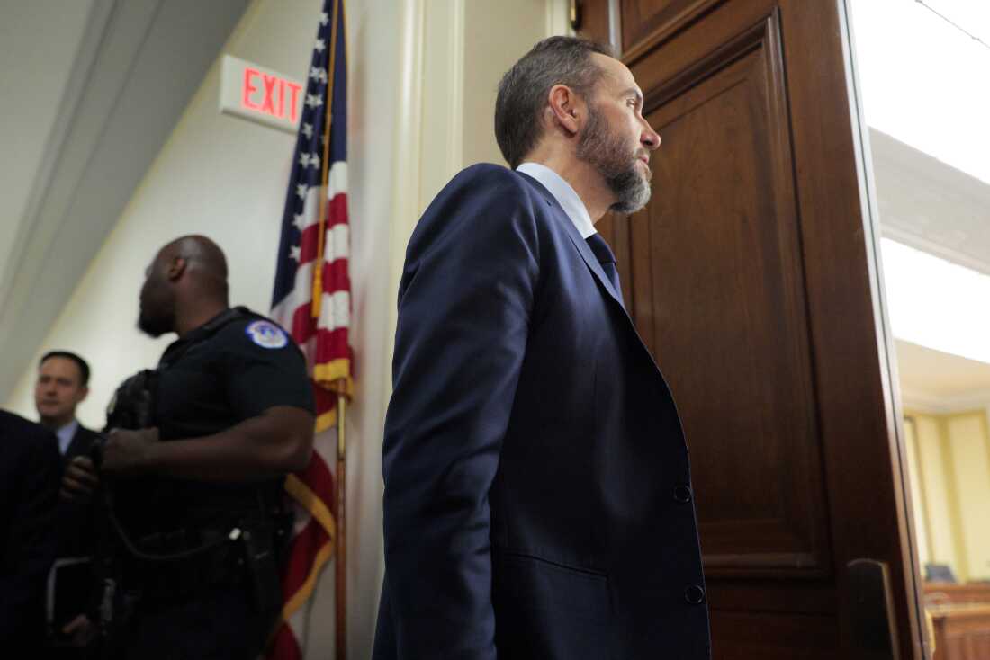 A bearded man wearing a suit approaches an open doorway with a U.S. flag to the side.