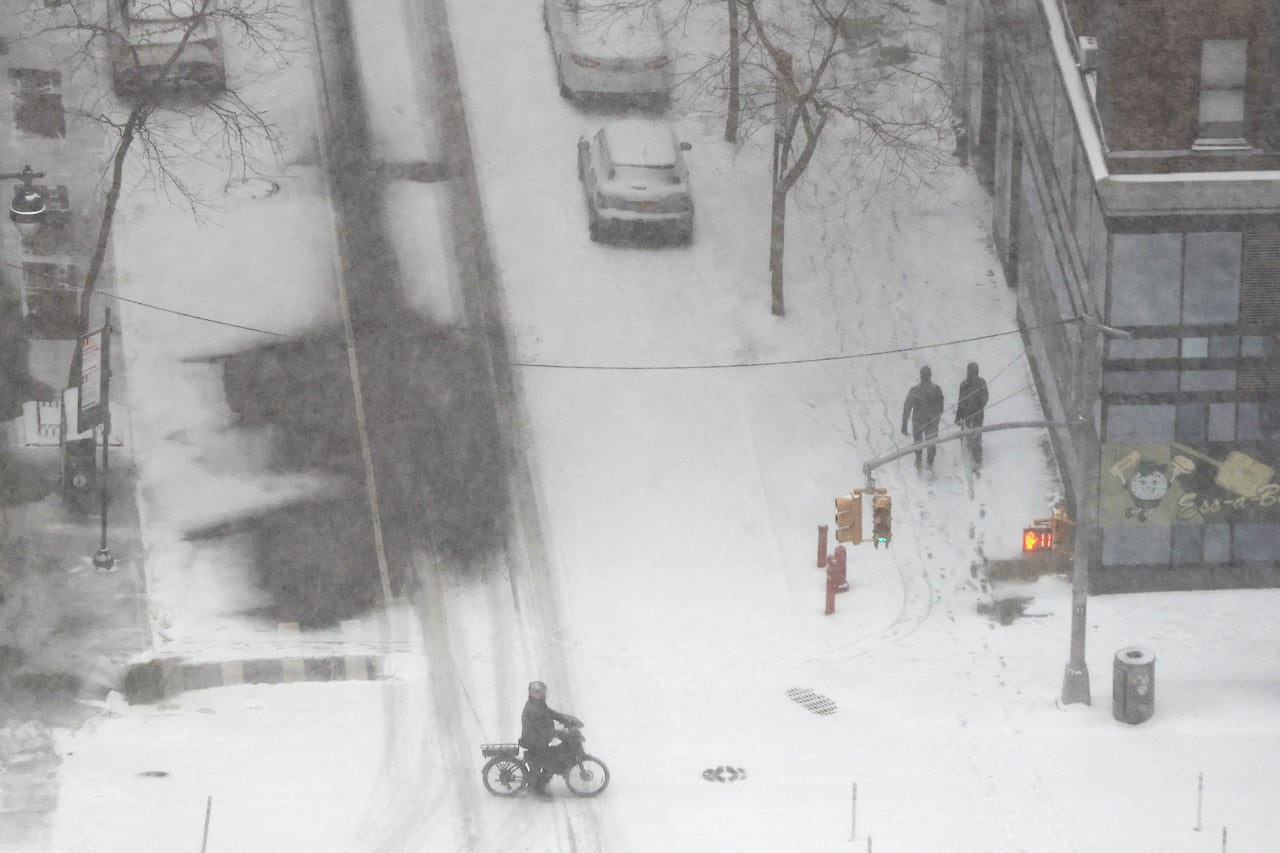 Snowy city streets, with pedestrians, a cyclist and traffic lights, are seen from above.
