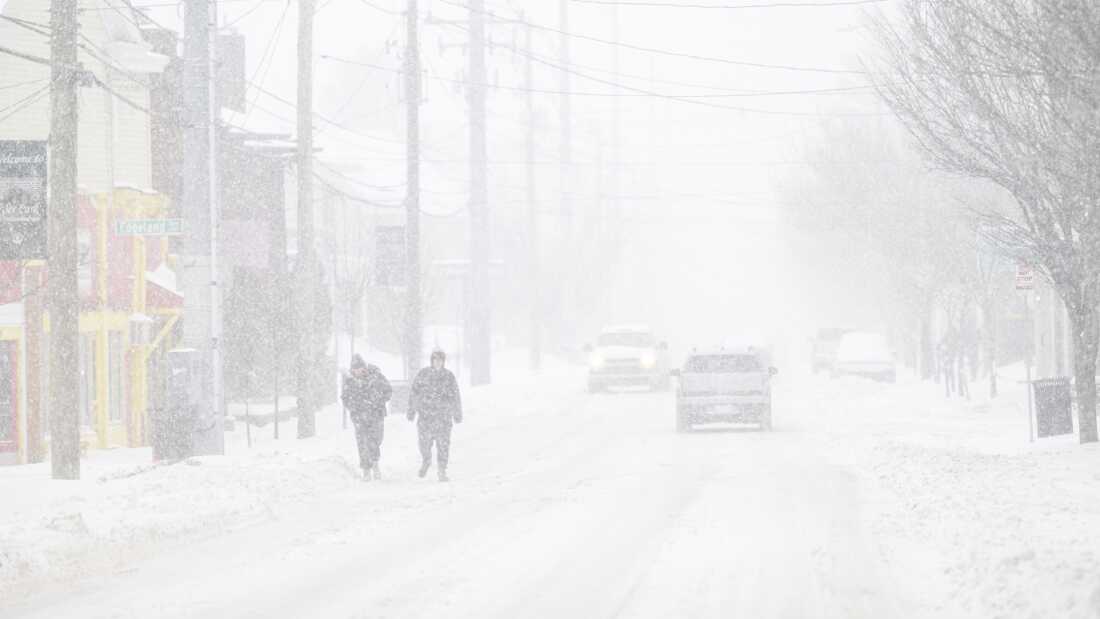 Two individuals walk along a street; they, the vehicles behind them, and the building next to them are blurred by snowfall.