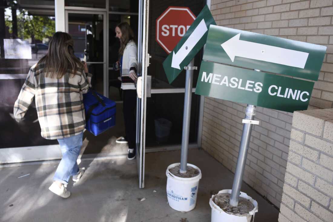A sign that says "Measles Clinic" is shown. Two people are entering the Andrews County Health Department in Texas.