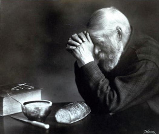 An elderly man with white hair and a beard sits at a table with his hands clasped in prayer over a loaf of bread, a bowl, a knife, and a closed book.