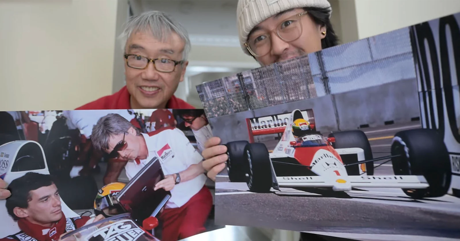 Two men smile at the camera while holding large photos of Formula 1 scenes, including a driver signing an autograph and a racing car on the track.