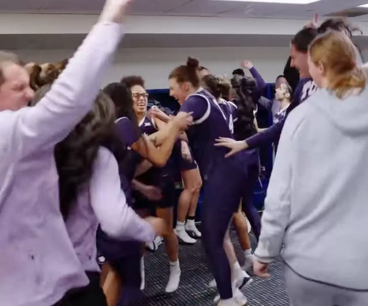 A group of individuals dressed in athletic uniforms and matching colors are celebrating and huddling together in a gymnasium. The individuals are engaging in gestures of joy and camaraderie, possibly in response to a recent achievement or victory.