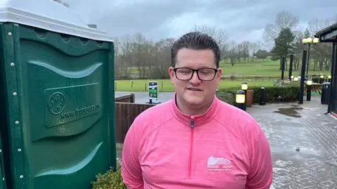 PHIL HARRISON/BBC Matthew Turner wears a salmon-coloured sports top and stands in front of a row of green portable toilets.