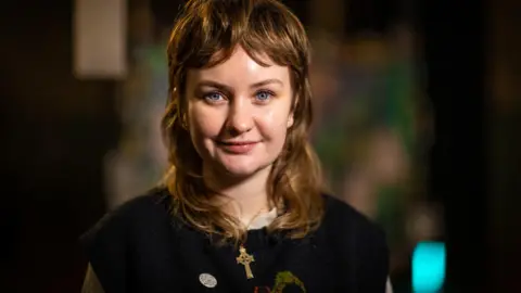BBC A woman wearing a dark knitted sweater with a cross necklace, standing indoors with soft, colourful, out‑of‑focus lights in the background.