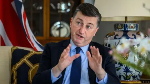 Getty Images Douglas Alexander in a dark suit, blue shirt and blue tie gesturing with his hands. He is sitting on a chair with a brown cushion on it and there is a cabinet, vase and union flag behind him 