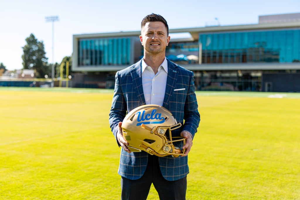 UCLA Football Head Coach Bob Chesney holding a gold helmet with "UCLA" written on it in blue.