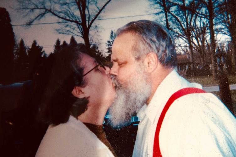 A woman wearing glasses kissing a man with a long gray beard.