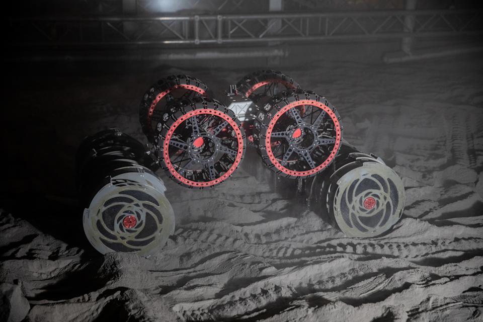 A wheeled robot navigates a sandy, dimly lit environment. Tracks in the sand indicate recent movement.