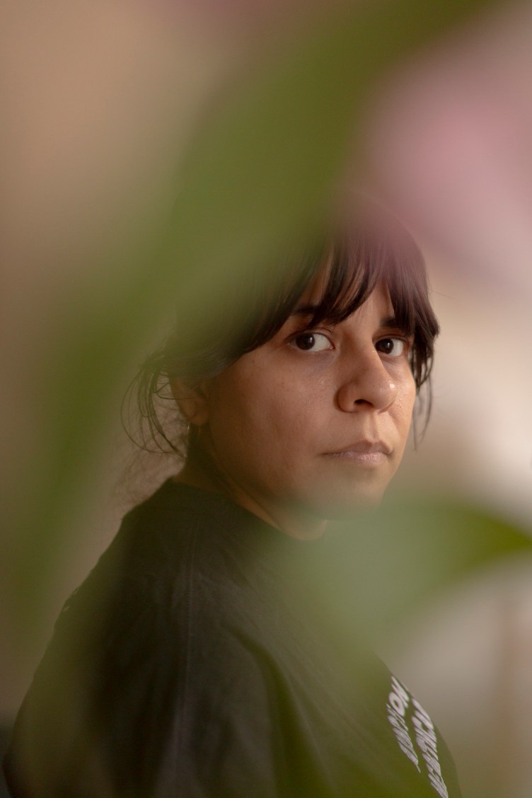 A woman with brown bangs and her hair pulled back wears a black T-shirt and small hoop earrings. She stares directly at the camera. Out-of-focus pink and green shapes slightly obscure her.