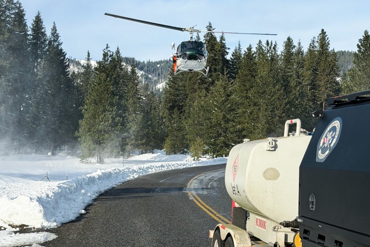 A helicopter flies above a street with a sheriff's truck parked on it and snow