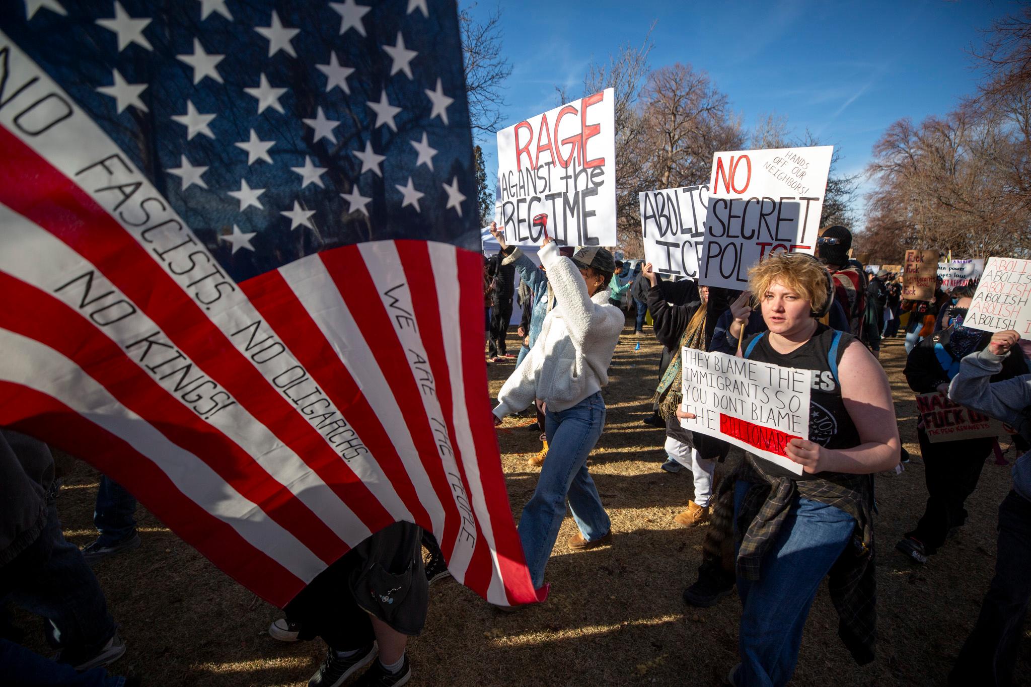 Half of the frame is filled by an American flag with words like "NO FASCISTS" written into the stripes. In the other half, a large crowd of people follow, most holding signs.