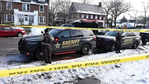 Getty Images Police tape is shown blocking off a snow-covered residential street. Two sheriff cars are in the foreground with officers standing in front of them. 