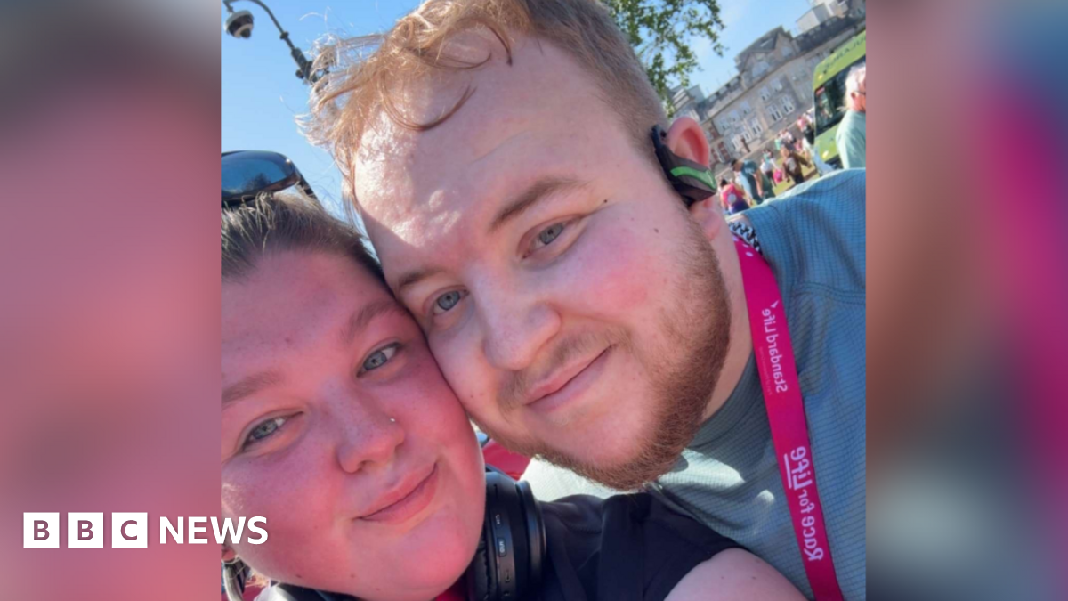 A close up selfie of Richard with his girlfriend, Sophie at the Race for Life running event. Sophie, on the left, has brown hair and wears sunglasses on the top of her head. Richard is standing next to Sophie with his cheek touching her cheek. He wears a green t-shirt, a pink medal, earphones and has short blonde hair.