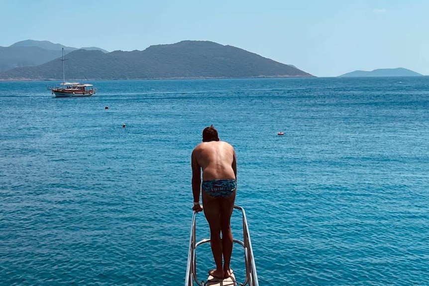 A man stands on the end of a boat, leaning on the rails and looking across the ocean with a mountain in the background.