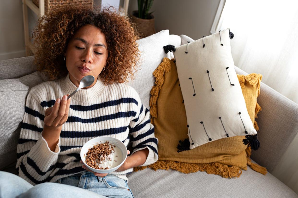 high angle view of young relaxed happy multiracial latina woman eating healthy granola and yogurt breakfast sitting on the couch at home cozy living room. copy space.