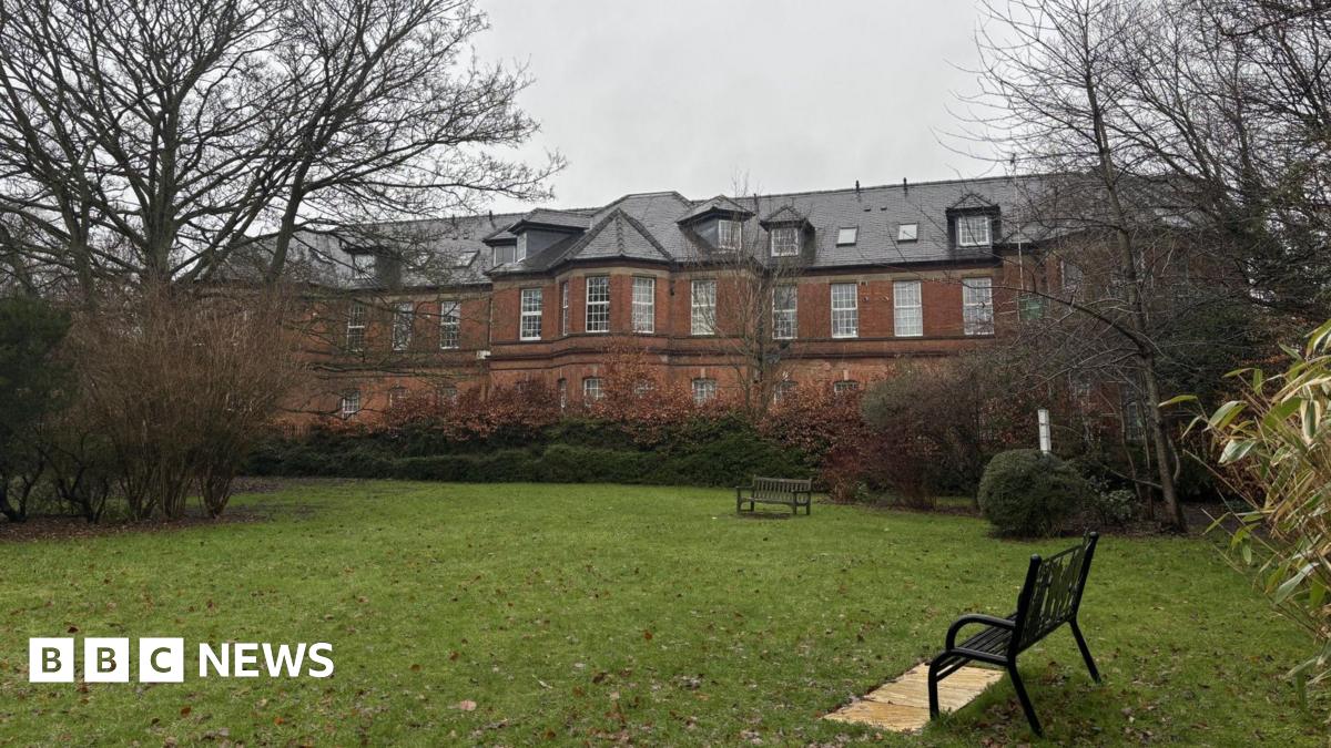 A red brick building with many windows behind a small park with benches, trees and hedges.