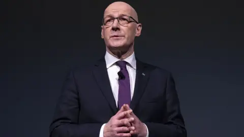 PA Media John Swinney, who is bald with glasses, is wearing a dark suit, white shirt and purple tie. He is standing with his hands clasped in front of him in front of a dark background. 