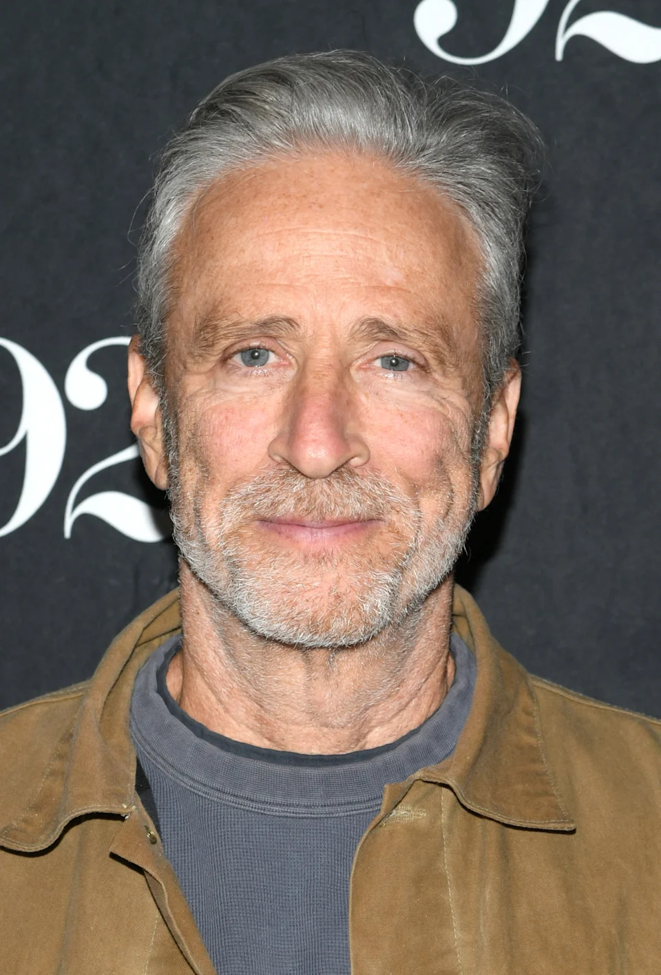 A person smiles at an event, wearing a casual jacket and t-shirt, standing against a branded backdrop