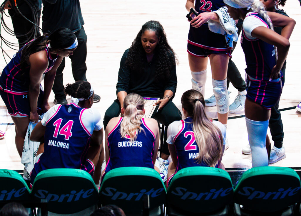 A coach addresses a group of players on the court. The coach is wearing black and the players are wearing blue and pink uniforms. 