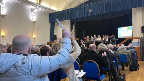 A group of people with their backs to the camera look towards a stage with other seated people