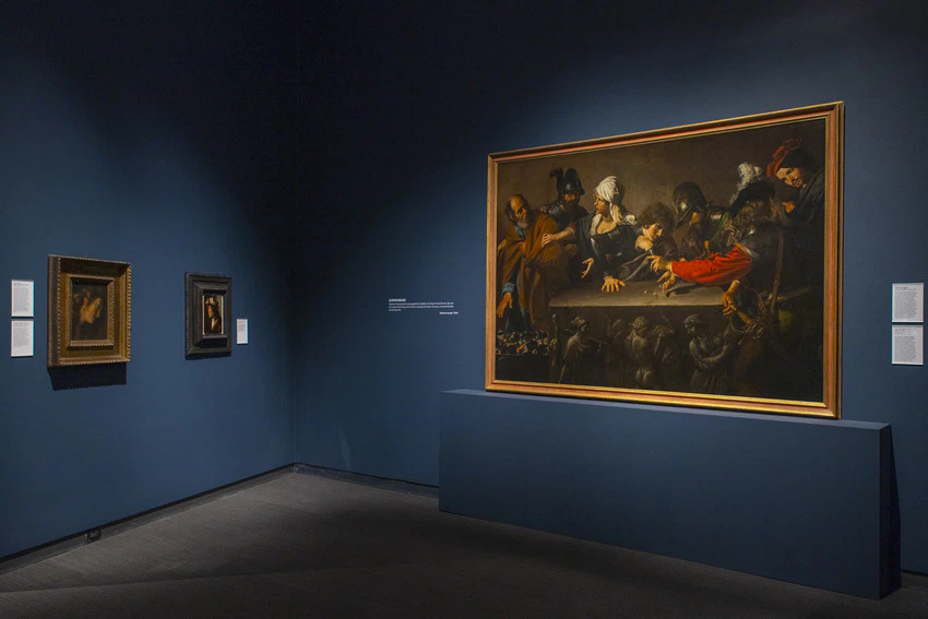 Art museum exhibit featuring a large historical painting on a blue wall alongside two smaller framed artworks.
