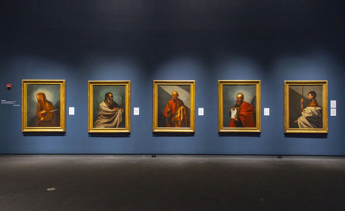 Five framed paintings of various figures in a museum gallery with a dark blue wall background.