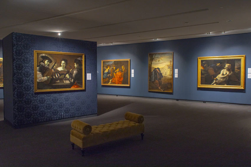 Art gallery interior with classical paintings on blue walls and a yellow bench in the center.