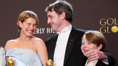 Reuters Jessie Buckley, Paul Mescal, and child actor Jacobi Jupe pose with the best film award at the Golden Globes.  Buckley, who has short blonde hair and an off-the-shoulder silver/pale blue gown, is smiling broadly with her eyes closed. Mescal has short, dark hair and is wearing a black suit and white shirt.  Jacobi Jupe has short blonde hair and is smiling. 
