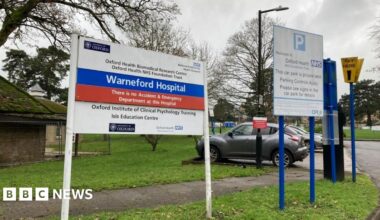 A sign outside a hospital saying Warneford Hospital. There's a carpark in the background and some autumnal trees.