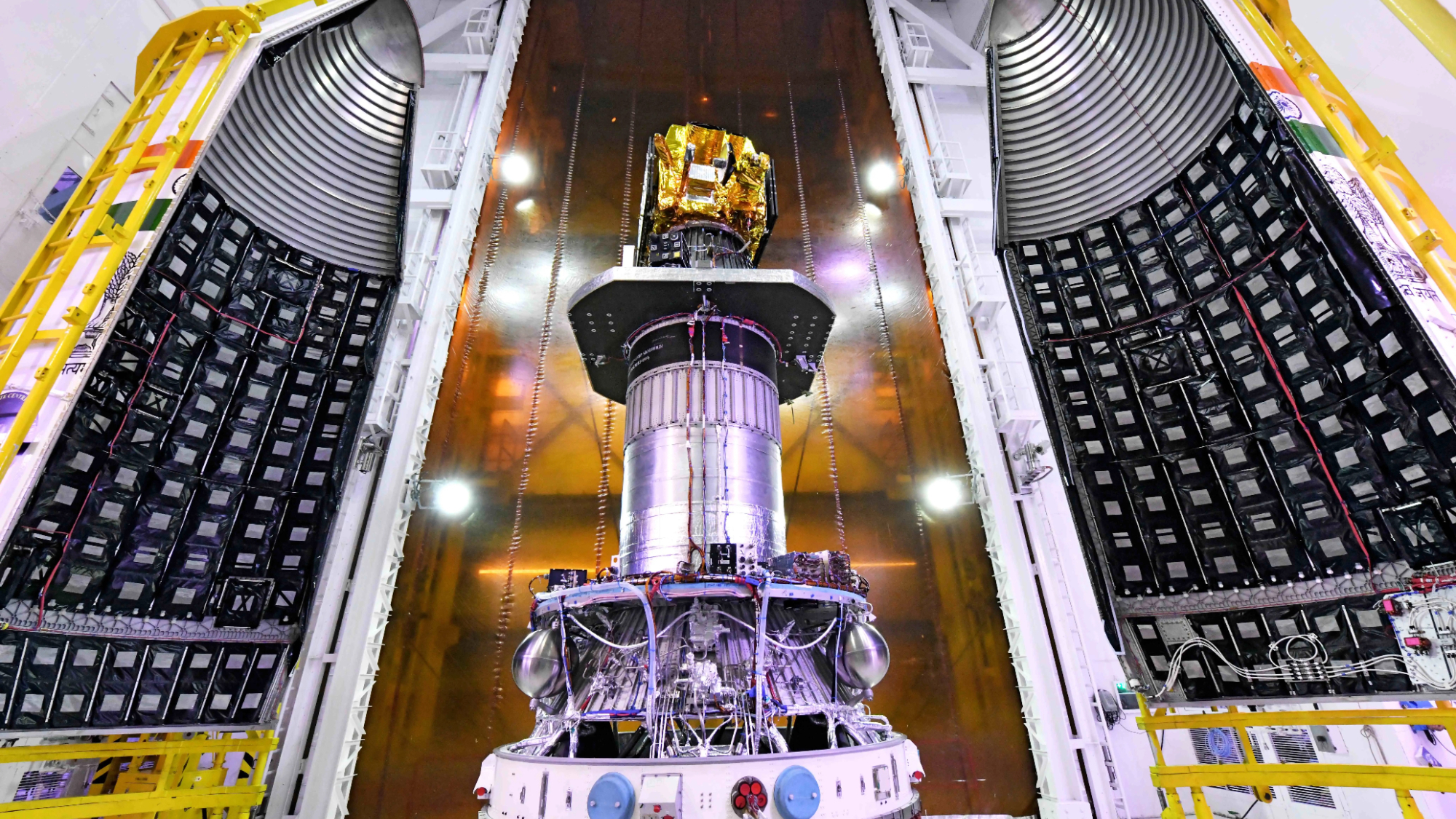 a boxy golden satellite is seen inside a large hangar being encapsulated into its payload fairing