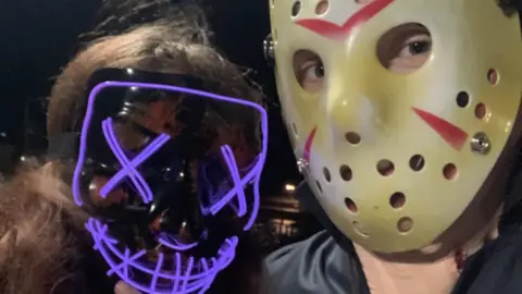 Ameila Relfe Ameila and Lewis Relfe wearing masks. She is on the left wearing a black mask with blue neon lights in the style popularised by the Purge movies. The eyes are crossed out and the smile has stitches across it. Lewis - on the right - is wearing a hockey mask featured in the Friday 13th 