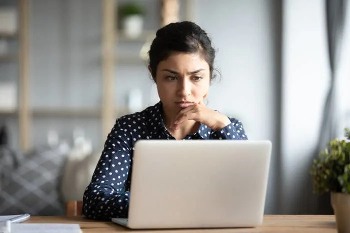 Person at desk with hand on chin, focused on a laptop screen. Background shows shelves and decor