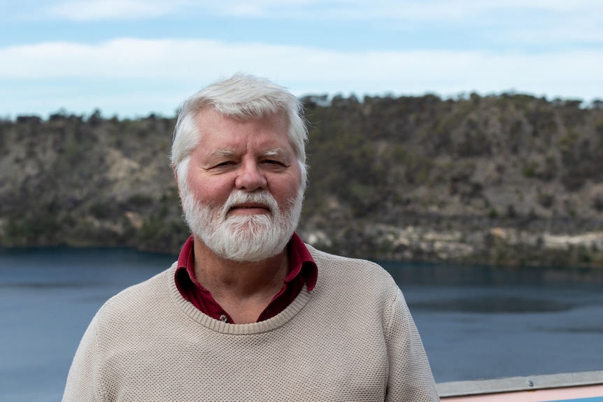 A man with white hair and a white beard standing in front of a lake.