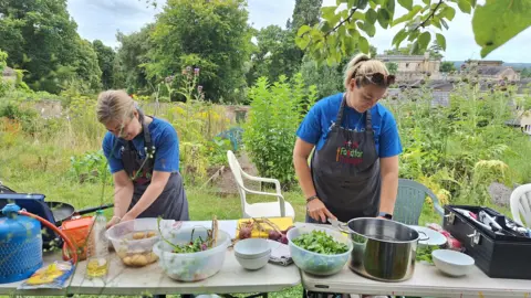 Bath Mind Two people cooking on two tables covered in foraged food. They are in a green garden on what seems like  a sunny day.