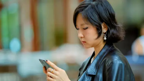 Getty Images Young woman in a leather jacket using a smartphone in an outdoor setting. Shanghai, China - stock photo