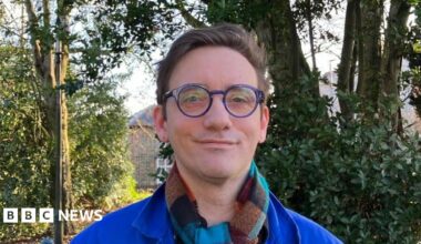 Shows a man in a blue jacket and checked scarf with round blue glasses standing in front of trees