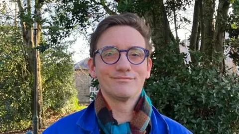 BBC Shows a man in a blue jacket and checked scarf with round blue glasses standing in front of trees