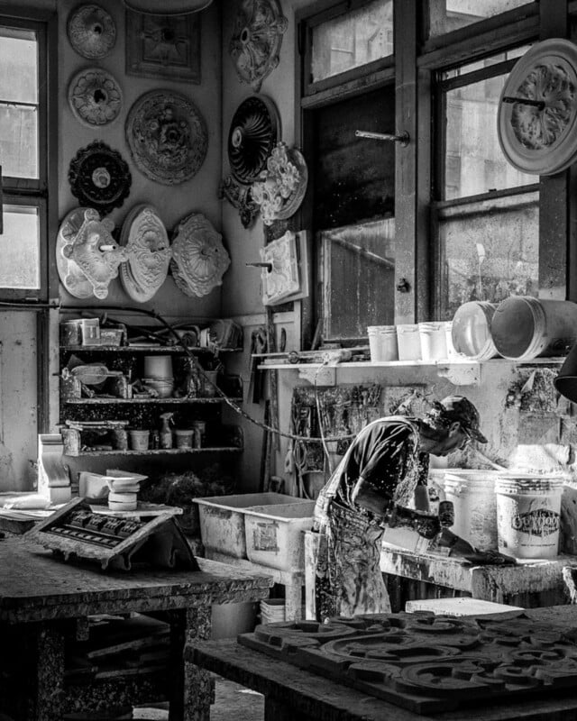 A person wearing overalls and a cap works with plaster in a sunlit workshop filled with decorative plaster molds, art tools, and buckets, with finished ornaments hanging on the walls. The photo is in black and white.