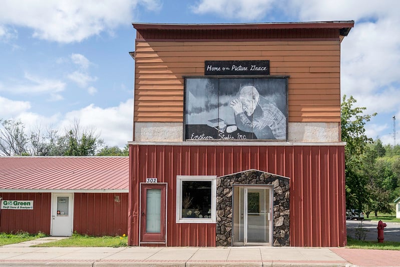 A two-story red and brown building with a mural of an elderly person painting, and the words "Home v. Prairie Dance" and "Einstein Studio Inc." above the entrance. Trees and another building are in the background.