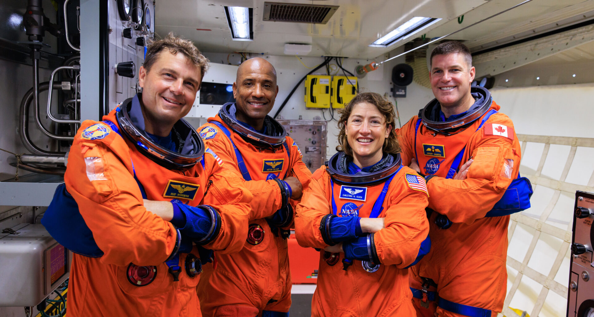 Four astronauts wearing an orange and blue spacesuit pose with their arms crossed inside of a facility.