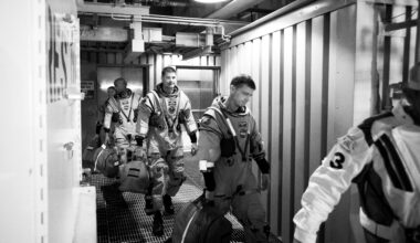 Four astronauts in spacesuits walk through a small corridor, following another person. The photo is in black and white.