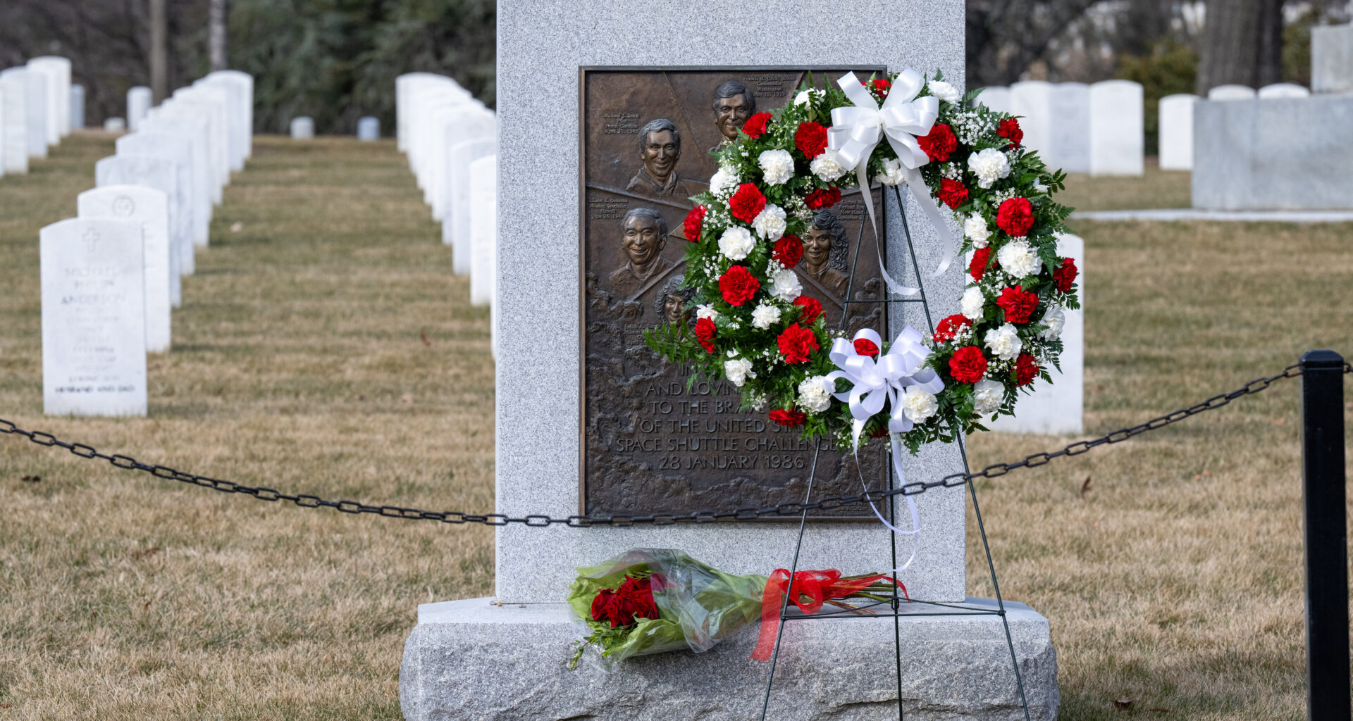A wreath with red and white flowers, green leaves, and white ribbon bows rests on a tripod stand with three thin metal legs. It stands in front of the Space Shuttle Challenger Memorial. The Memorial has two parts: A polished gray stone headstone with a bronze embossed plaque on it, resting on a small platform of rough gray stone. The plaque has the likeness of Challenger and the crew on it. Behind the memorial, we can see rows of white stone headstones.