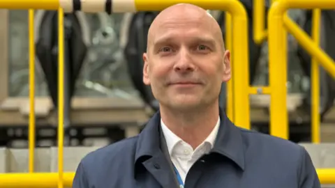A man with a white shirt and a blue coat is looking at the camera. Yellow railings are behind him.