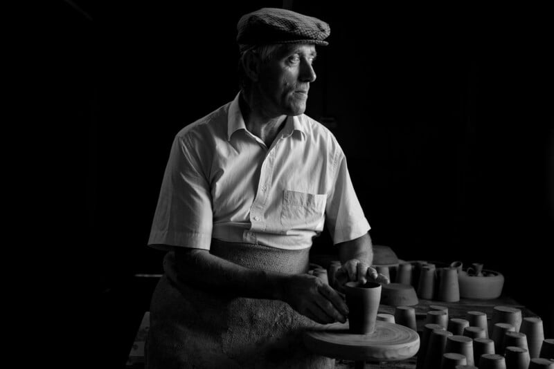 A man wearing a flat cap and apron shapes a clay pot on a pottery wheel, surrounded by other clay items, in a dimly lit workshop.